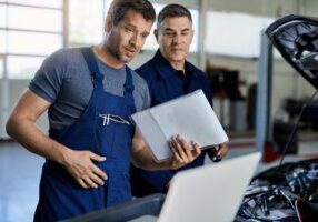 Auto repairman and his coworker using laptop while analyzing car's performance in a workshop.